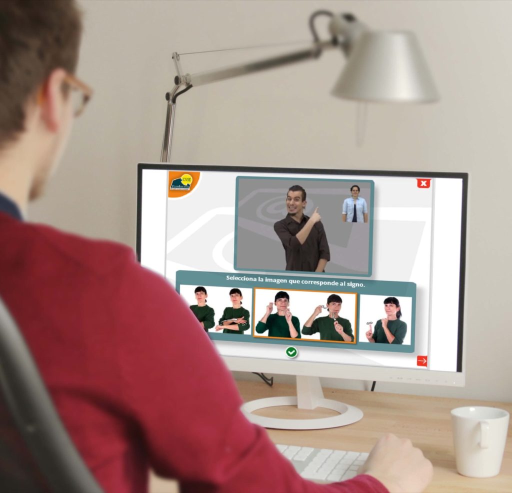 Chico de espaldas, jersey rojo y gafas, mira una pantalla de ordenador con imágenes de clases de lengua de signos, hay una taza blanca en la mesa