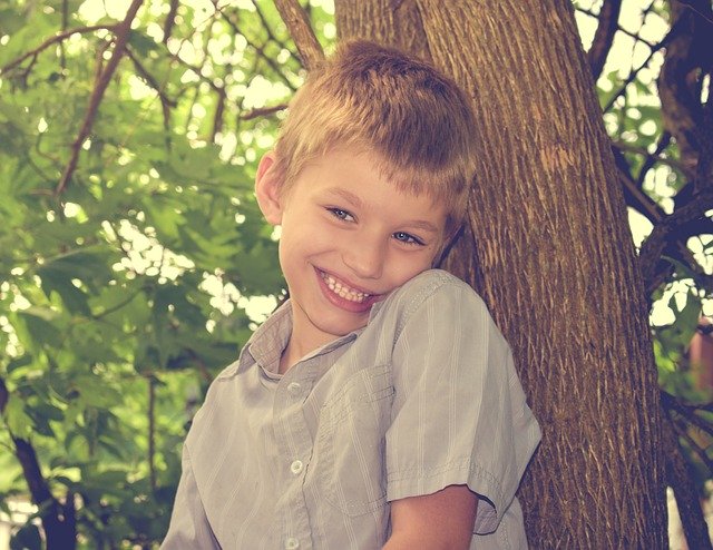 Niño de aproximadamente diez a doce años, pelo rubio y corto, sonrisa amplia, camisa color gris y rayas verticales blancas, apoyado en un árbol